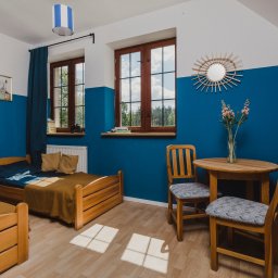 KPCEEiS GRZEGORZ WĄS - Pokój w ośrodku wypoczynkowym z dwoma drewnianymi łóżkami, okrągłym stolikiem i krzesłami, udekorowany kwiatami w wazonie i lustrem w kształcie słońca na ścianie pomalowanej na biało i niebiesko.