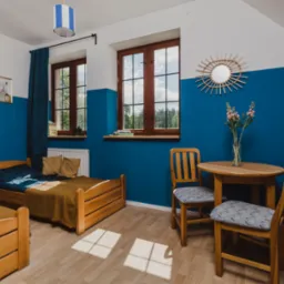 Pokój w ośrodku wypoczynkowym z dwoma drewnianymi łóżkami, okrągłym stolikiem i krzesłami, udekorowany kwiatami w wazonie i lustrem w kształcie słońca na ścianie pomalowanej na biało i niebiesko.