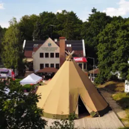 Widok z góry na duży namiot tipi przed budynkiem Kujawsko-Pomorskiego Centrum Edukacji Ekologicznej, w tle las i food truck.