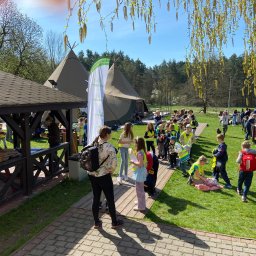 KPCEEiS GRZEGORZ WĄS - Grupa dzieci w odblaskowych kamizelkach na zielonym terenie z namiotami tipi w tle, widok z pod drewnianej altany w słoneczny dzień.