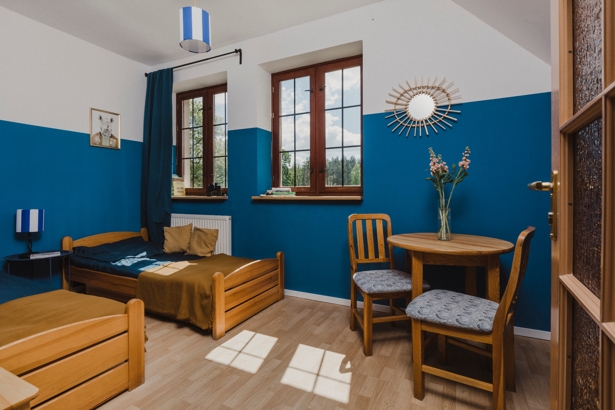 Pokój w ośrodku wypoczynkowym z dwoma drewnianymi łóżkami, okrągłym stolikiem i krzesłami, udekorowany kwiatami w wazonie i lustrem w kształcie słońca na ścianie pomalowanej na biało i niebiesko.