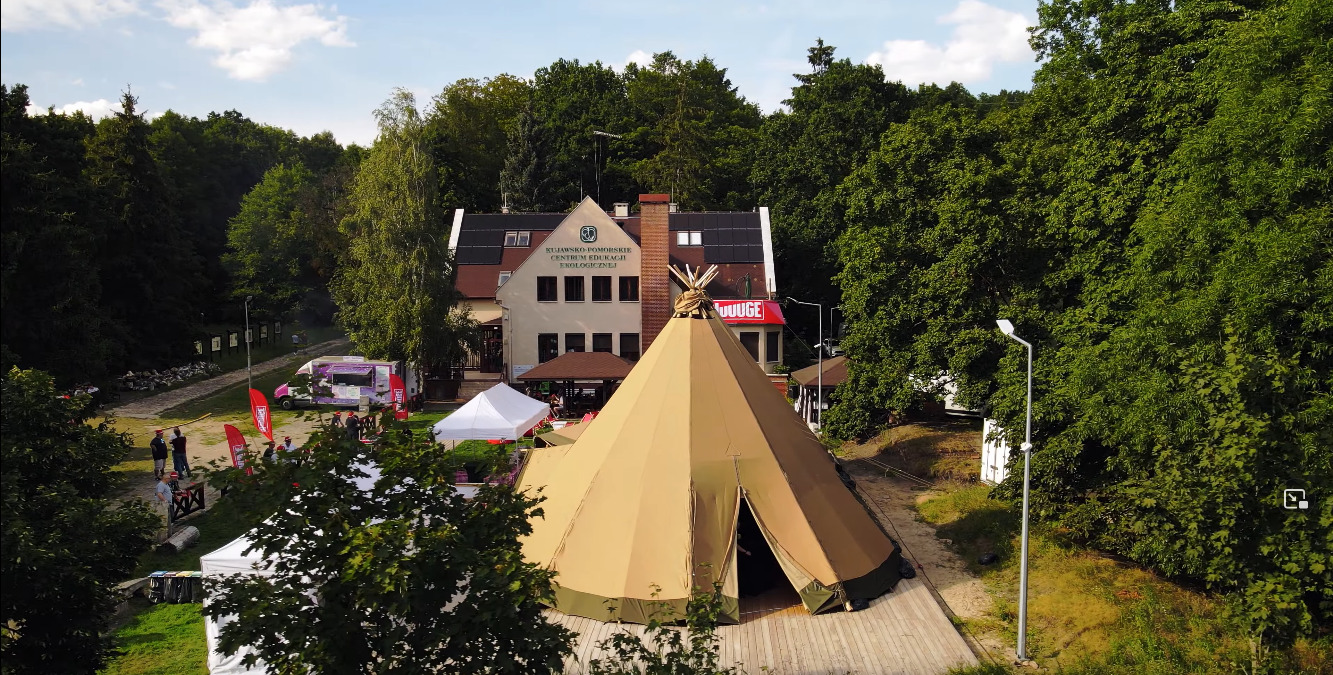 Widok z góry na duży namiot tipi przed budynkiem Kujawsko-Pomorskiego Centrum Edukacji Ekologicznej, w tle las i food truck.