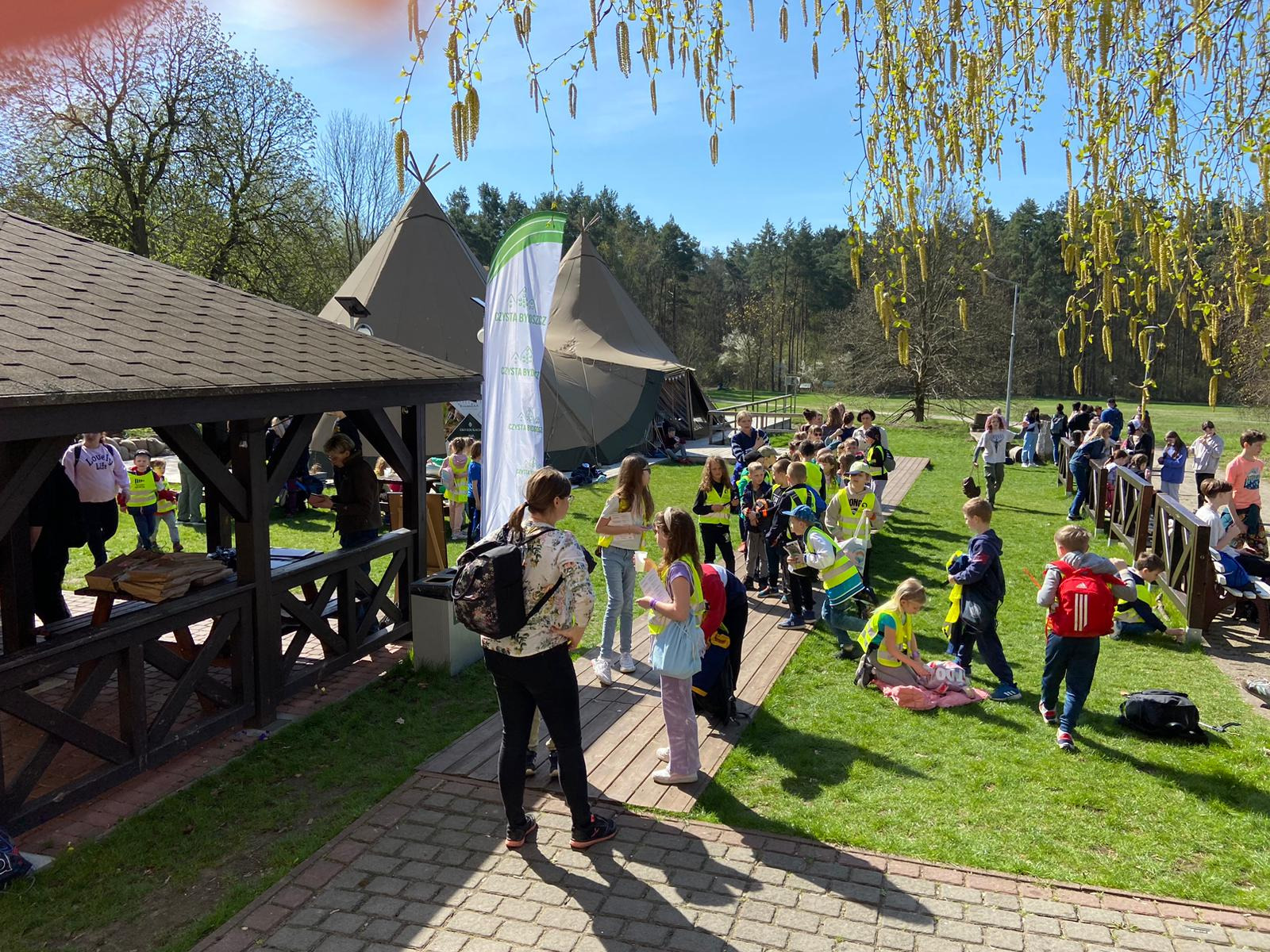 Grupa dzieci w odblaskowych kamizelkach na zielonym terenie z namiotami tipi w tle, widok z pod drewnianej altany w słoneczny dzień.