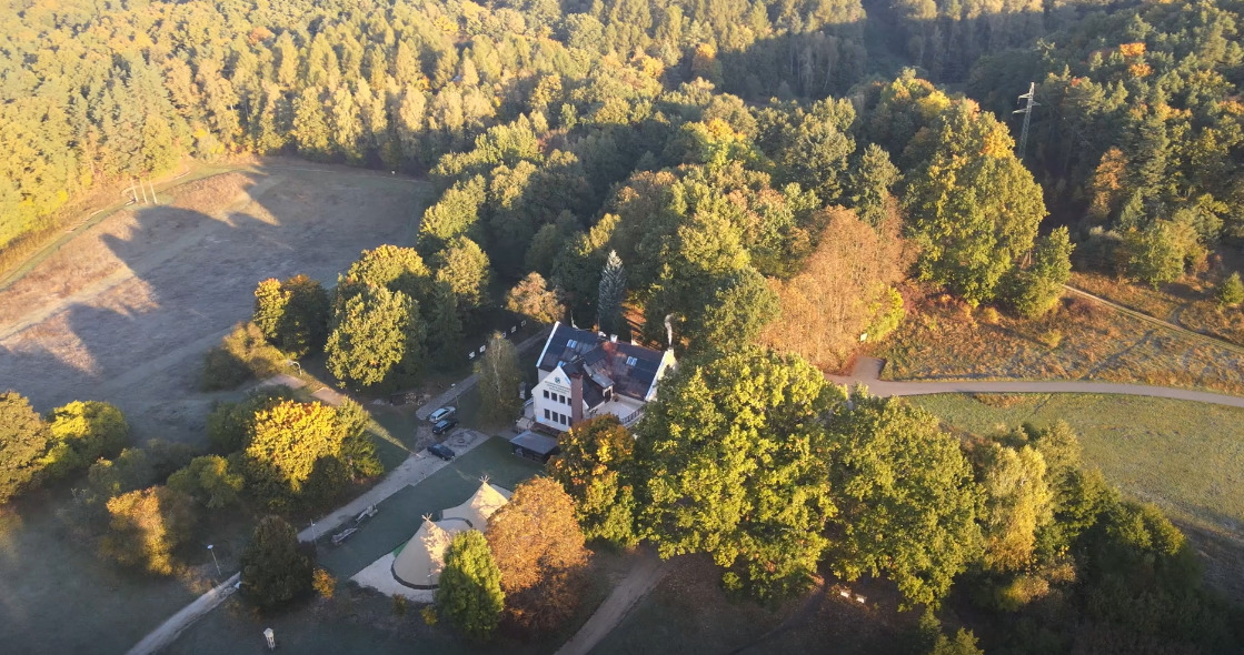 Widok z lotu ptaka na ośrodek wypoczynkowy otoczony lasem, z wigwamami i budynkiem głównym z jasną elewacją i ciemnym dachem, wczesnym rankiem, w jesiennej scenerii.