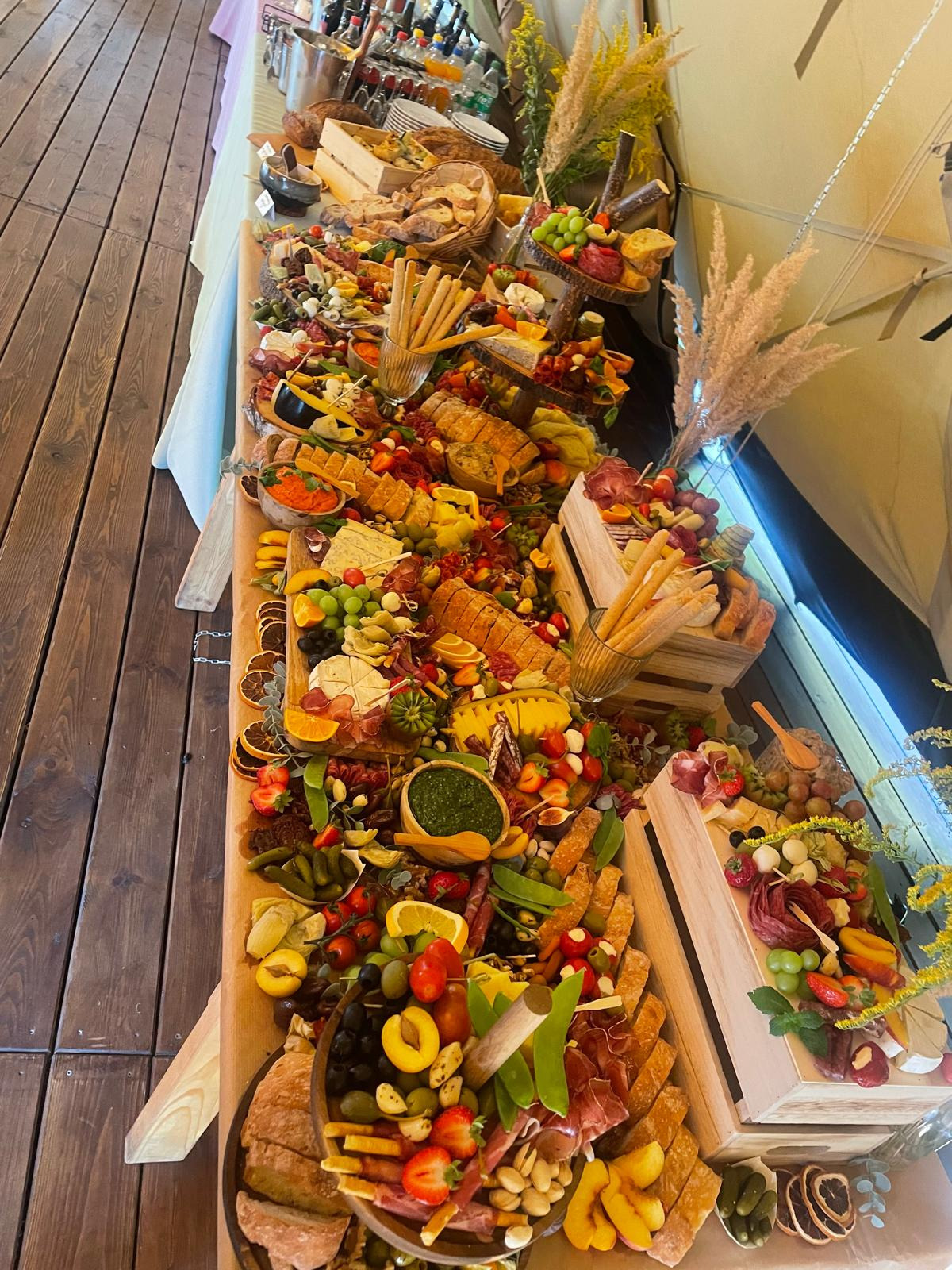 Bogato zastawiony stół przekąskami: sery, owoce, pieczywo, wędliny, pesto w drewnianych miseczkach, całość udekorowana suszonymi trawami.