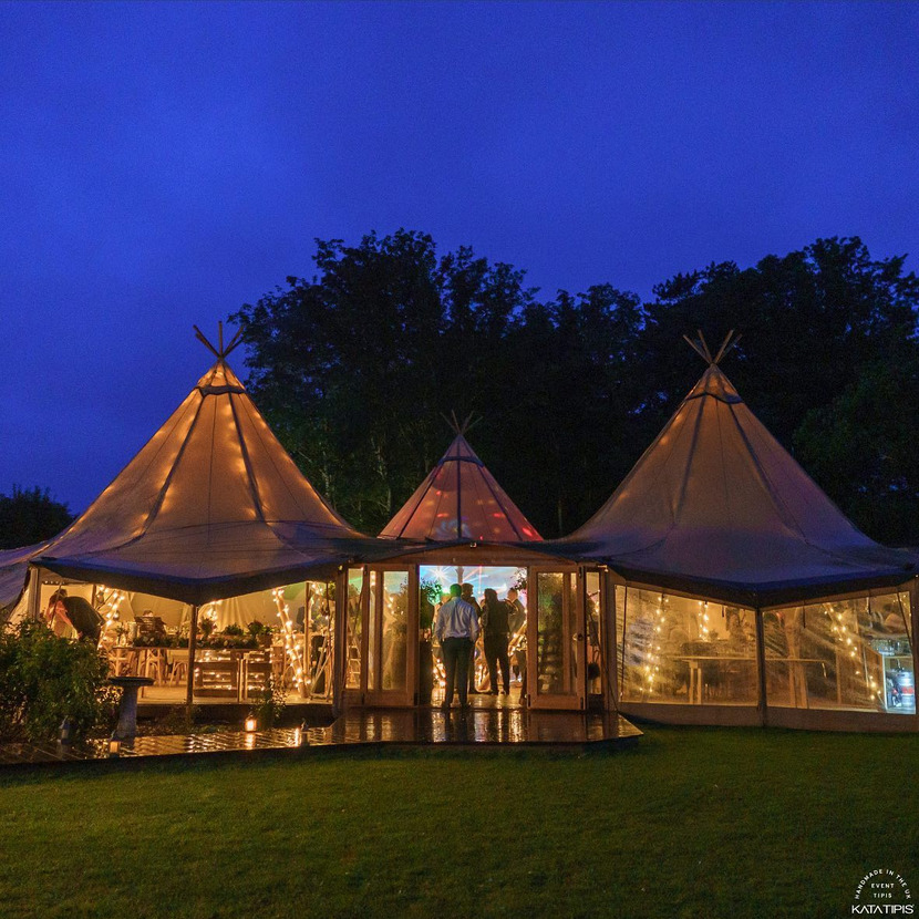 Oświetlony namiot tipi z przezroczystymi ścianami, wewnątrz widoczni ludzie i dekoracje świetlne, na zewnątrz trawnik i ciemne niebo.
