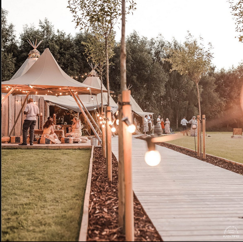 Uroczystość plenerowa w namiotach tipi oświetlonych girlandami, drewniany chodnik prowadzi do przestrzeni z gośćmi.