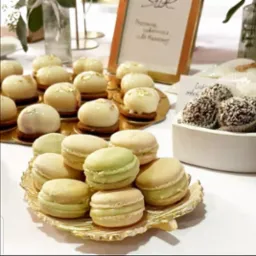 Elegancko podane desery: makaroniki na złotej paterze w kształcie liścia, mini-ciastka z białą polewą i złotymi płatkami oraz kulki wiórkowe w białym naczyniu.