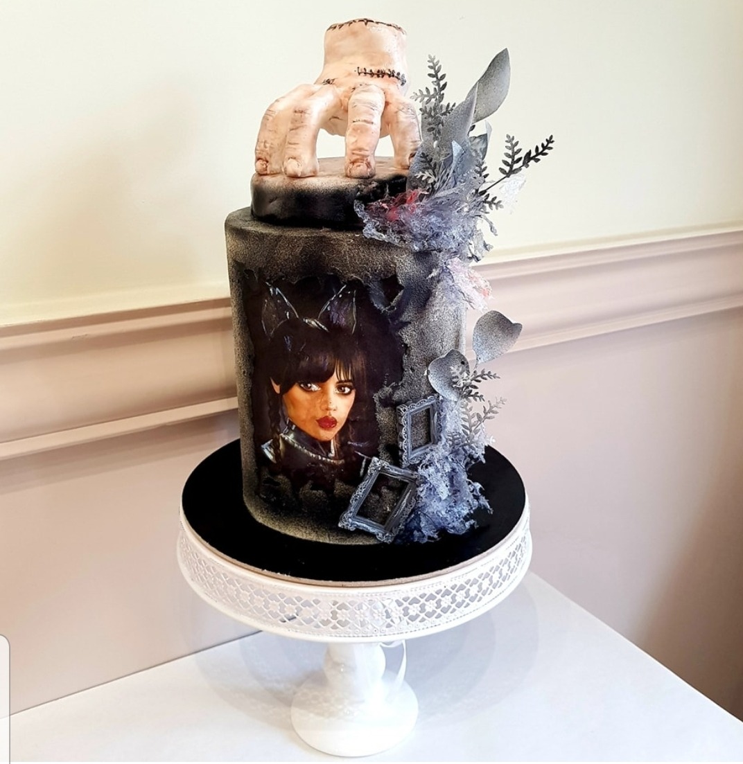 Dwupiętrowy tort z motywem Wednesday Addams: na górze ręka z bliznami, na boku portret bohaterki i srebrne dekoracje.