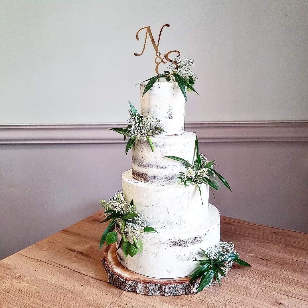 Czteropiętrowy tort weselny w stylu rustykalnym, z białym kremem, dekorowany zielonymi liśćmi i drobnymi białymi kwiatami, umieszczony na drewnianym plastrze pnia, z topperem w kolorze złotym...
