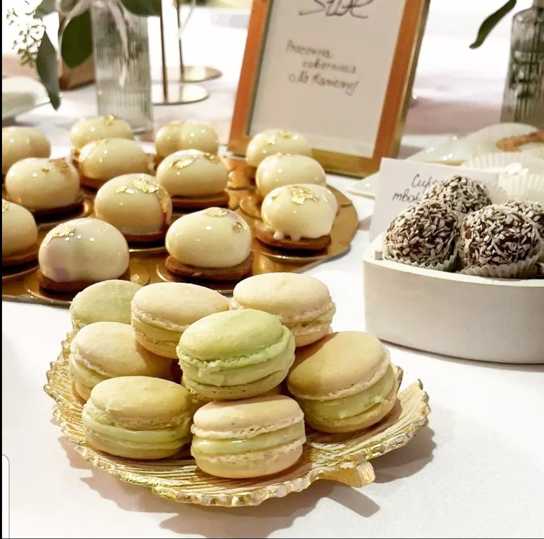 Elegancko podane desery: makaroniki na złotej paterze w kształcie liścia, mini-ciastka z białą polewą i złotymi płatkami oraz kulki wiórkowe w białym naczyniu.