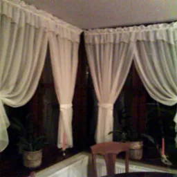 Delikatne, białe, zwiewne zasłony z falbanką upięte w oknach, z widocznymi doniczkami z kwiatami na parapecie i fragmentem krzesła w kadrze.