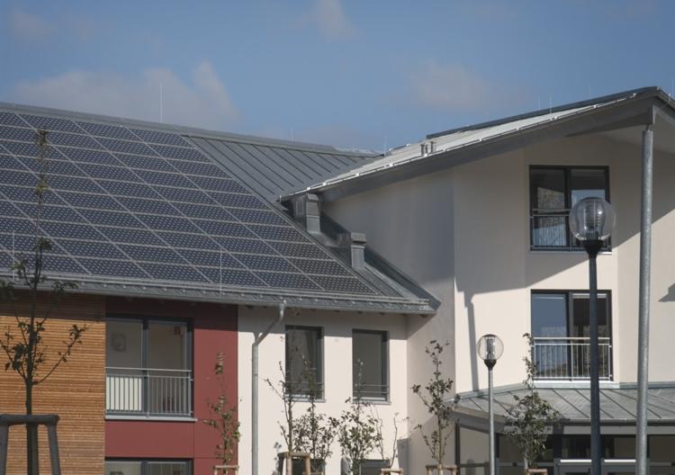 Panele fotowoltaiczne na dachu budynku mieszkalnego z metalowym dachem, widoczne okna z balkonami i lampy uliczne.