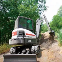 Mini koparka Bobcat E32 w trakcie prac ziemnych, z uniesionym ramieniem i łyżką nabierającą ziemię na tle zieleni.