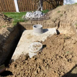 Betonowy zbiornik, prawdopodobnie szambo, umieszczony w wykopie ziemnym z widocznymi betonowymi elementami i pryzmą kamieni w tle, pod drewnianym płotem.