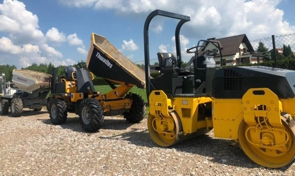 Żółty walec drogowy oraz wywrotki Thwaites na żwirowym placu, w tle dom jednorodzinny i ogrodzenie z siatki.