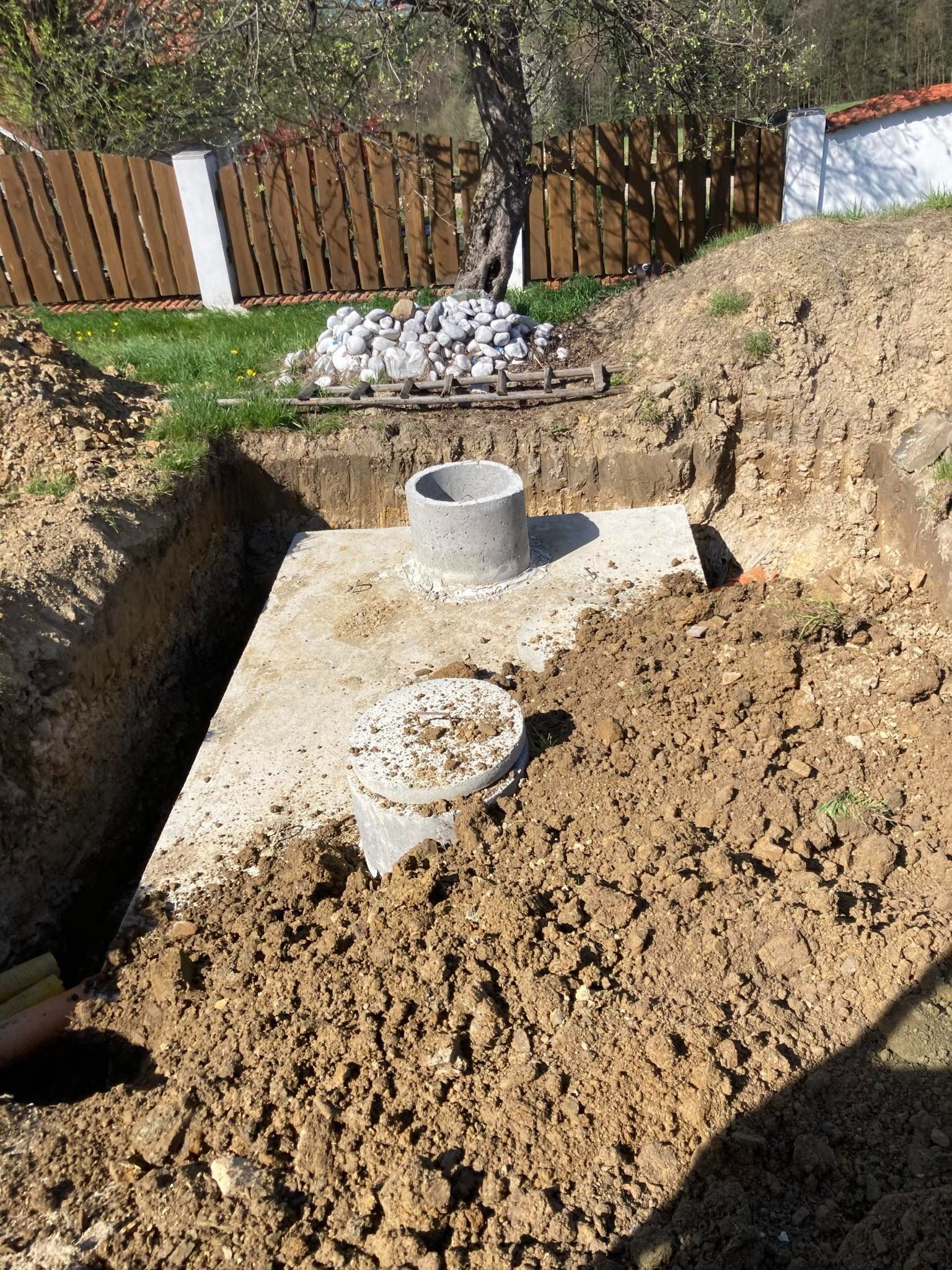 Betonowy zbiornik, prawdopodobnie szambo, umieszczony w wykopie ziemnym z widocznymi betonowymi elementami i pryzmą kamieni w tle, pod drewnianym płotem.