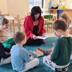 Niepubliczne Przedszkole i Żłobek Jezioraczek 3 - Grupa dzieci siedzi na niebieskim dywanie w kręgu, otoczona przez dwie kobiety, jedna czyta im z kartki, a przed nimi leżą małe doniczki w pudełku.