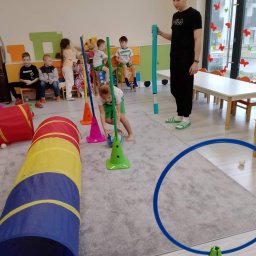 Niepubliczne Przedszkole i Żłobek Jezioraczek 3 - Wnętrze sali zabaw: dziewczynka w zielonym stroju prowadzi piłkę między zielonymi pachołkami, w tle dzieci siedzą na krzesłach, a mężczyzna w czarnym dresie trzyma niebieską rurę.