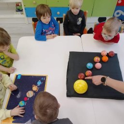 Niepubliczne Przedszkole i Żłobek Jezioraczek 3 - Grupa dzieci w żłobku lub przedszkolu siedzi przy stole, ucząc się o układzie słonecznym za pomocą modelu planet i planszy edukacyjnej, wskazując palcem na słońce.
