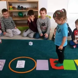 Dzieci bawią się w grę planszową z kolorowymi polami na dywanie, rzucają kostką i przechodzą przez obręcz z napisem META.