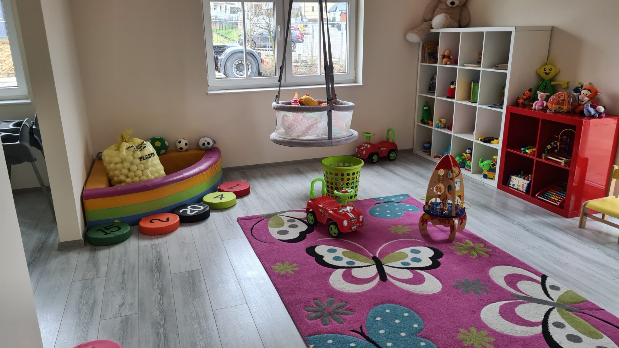 Pokój zabaw dla dzieci z basenem wypełnionym piłeczkami, wiszącym siedziskiem, regałem z zabawkami i dywanem w motyle.