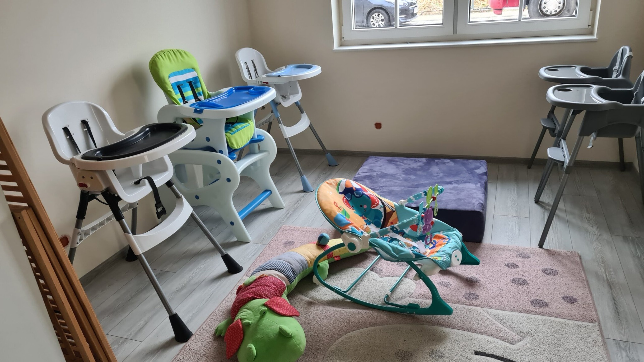 Pokój zabaw dla dzieci z kilkoma krzesełkami do karmienia, leżaczkiem-bujaczkiem i pluszową gąsienicą na dywanie.