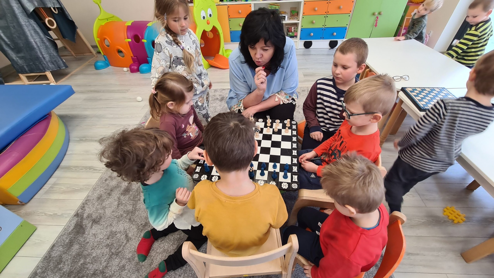 Grupa dzieci siedzi wokół szachownicy z opiekunką, która uważnie obserwuje grę; kolorowe meble i zabawki w tle.