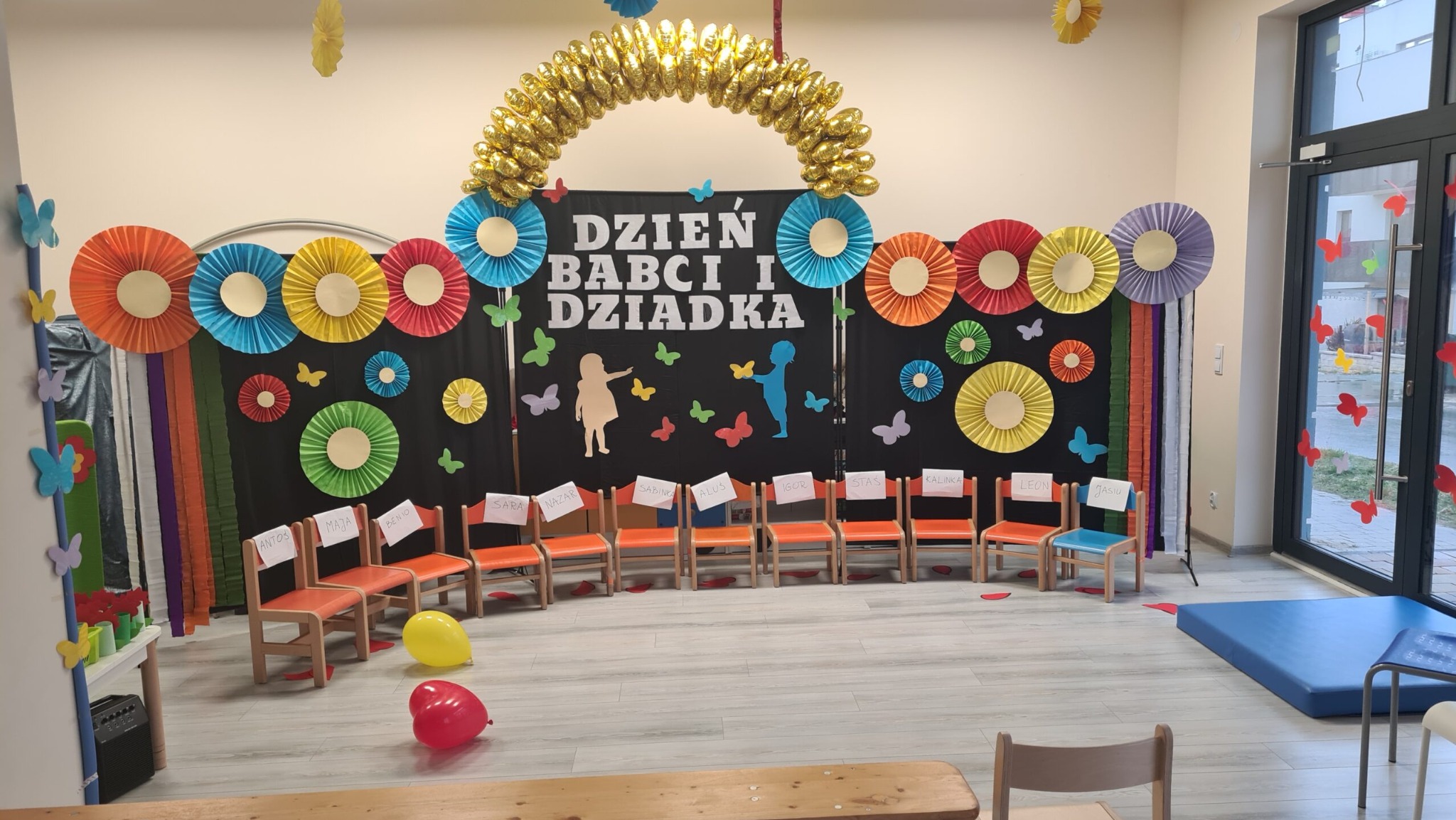Dekoracja sali z okazji Dnia Babci i Dziadka: czarne tło z kolorowymi papierowymi rozetami, złotym łukiem z balonów, sylwetkami dzieci i motylkami, krzesła z imionami dzieci, dwa balony na podłodze.