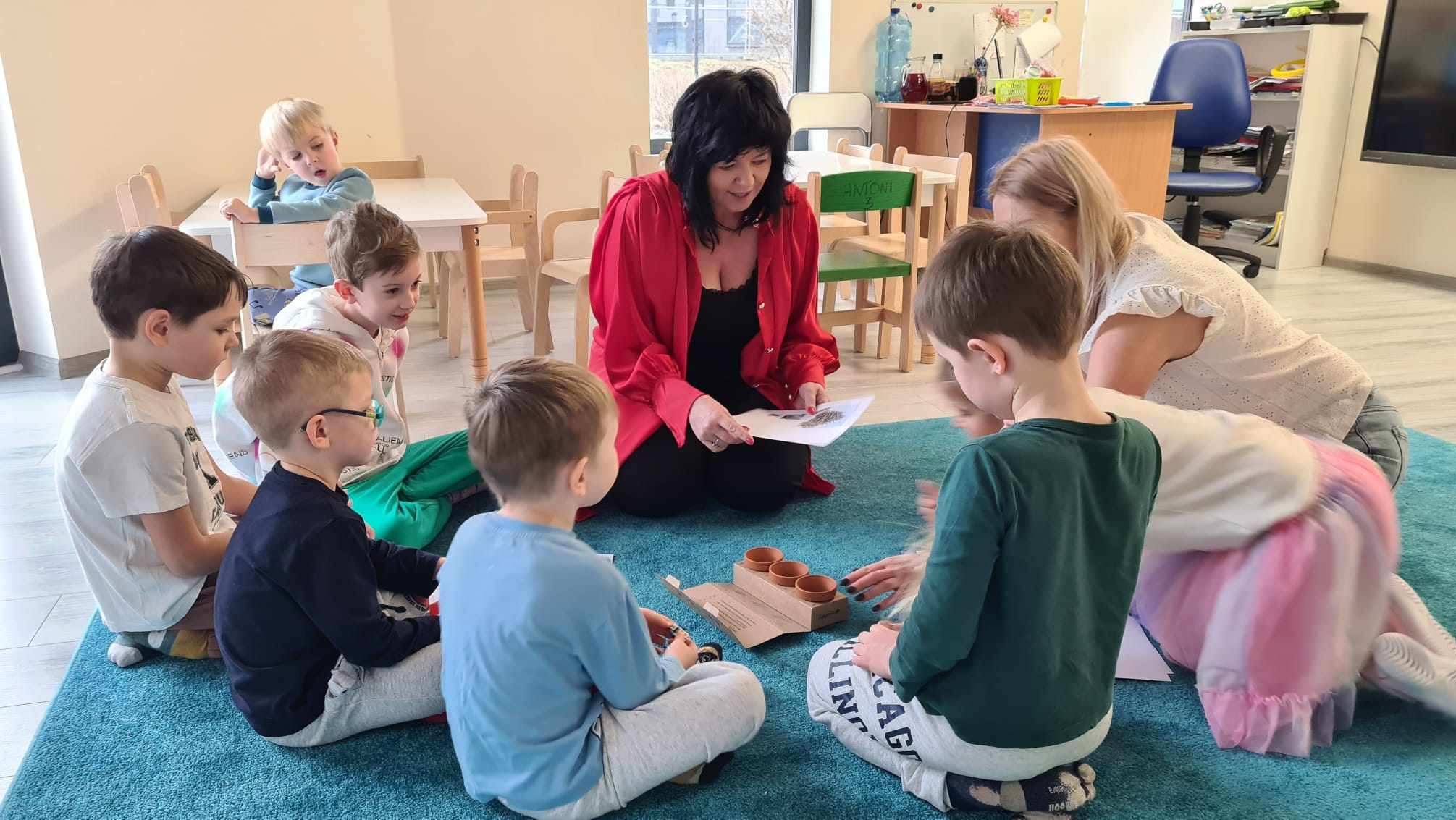 Grupa dzieci siedzi na niebieskim dywanie w kręgu, otoczona przez dwie kobiety, jedna czyta im z kartki, a przed nimi leżą małe doniczki w pudełku.