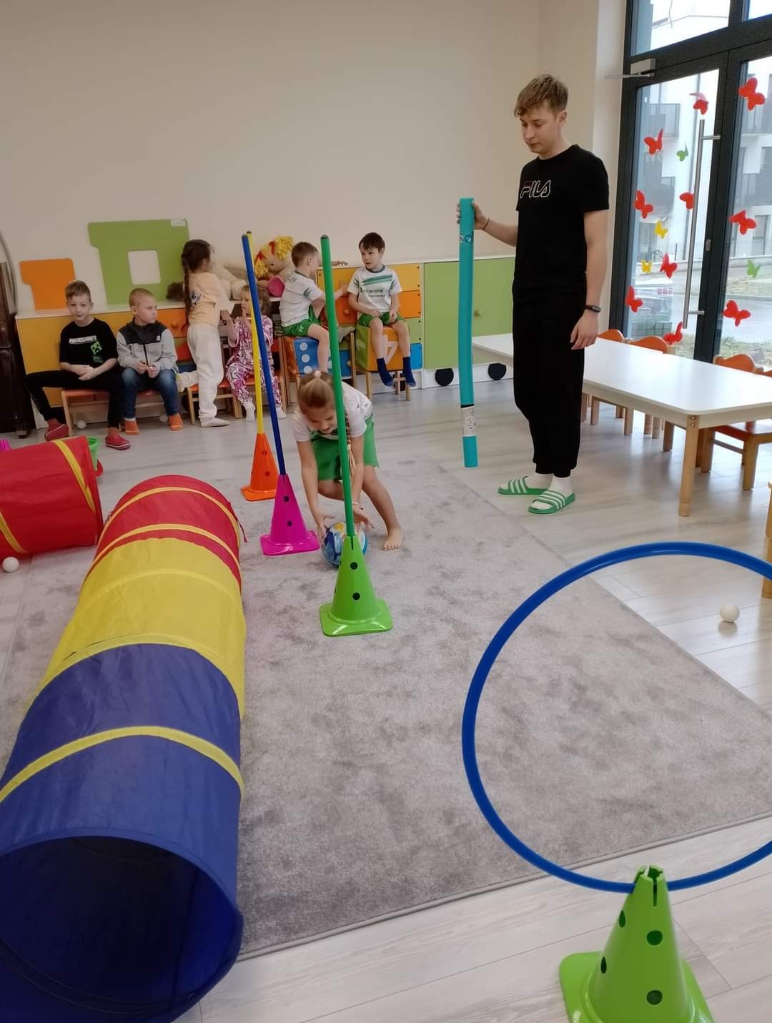 Wnętrze sali zabaw: dziewczynka w zielonym stroju prowadzi piłkę między zielonymi pachołkami, w tle dzieci siedzą na krzesłach, a mężczyzna w czarnym dresie trzyma niebieską rurę.