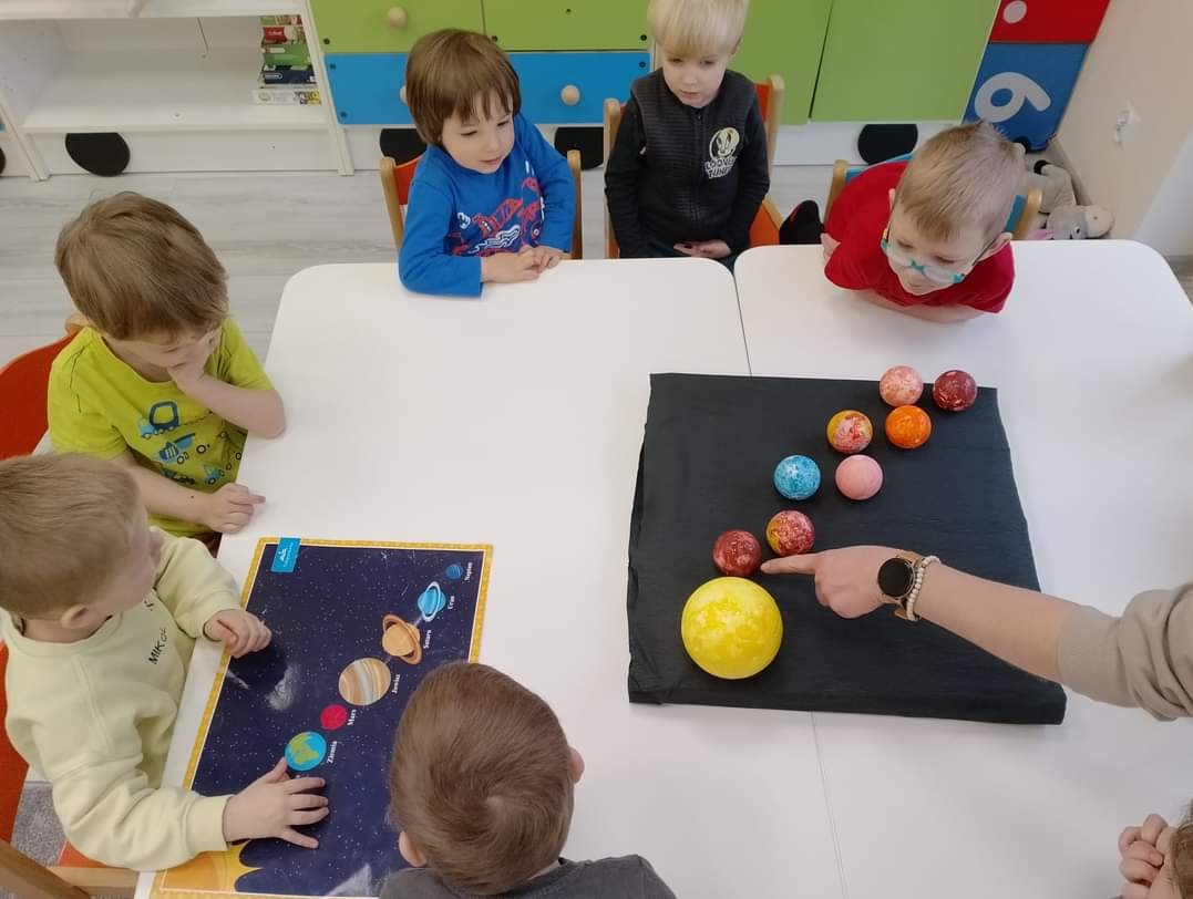 Grupa dzieci w żłobku lub przedszkolu siedzi przy stole, ucząc się o układzie słonecznym za pomocą modelu planet i planszy edukacyjnej, wskazując palcem na słońce.