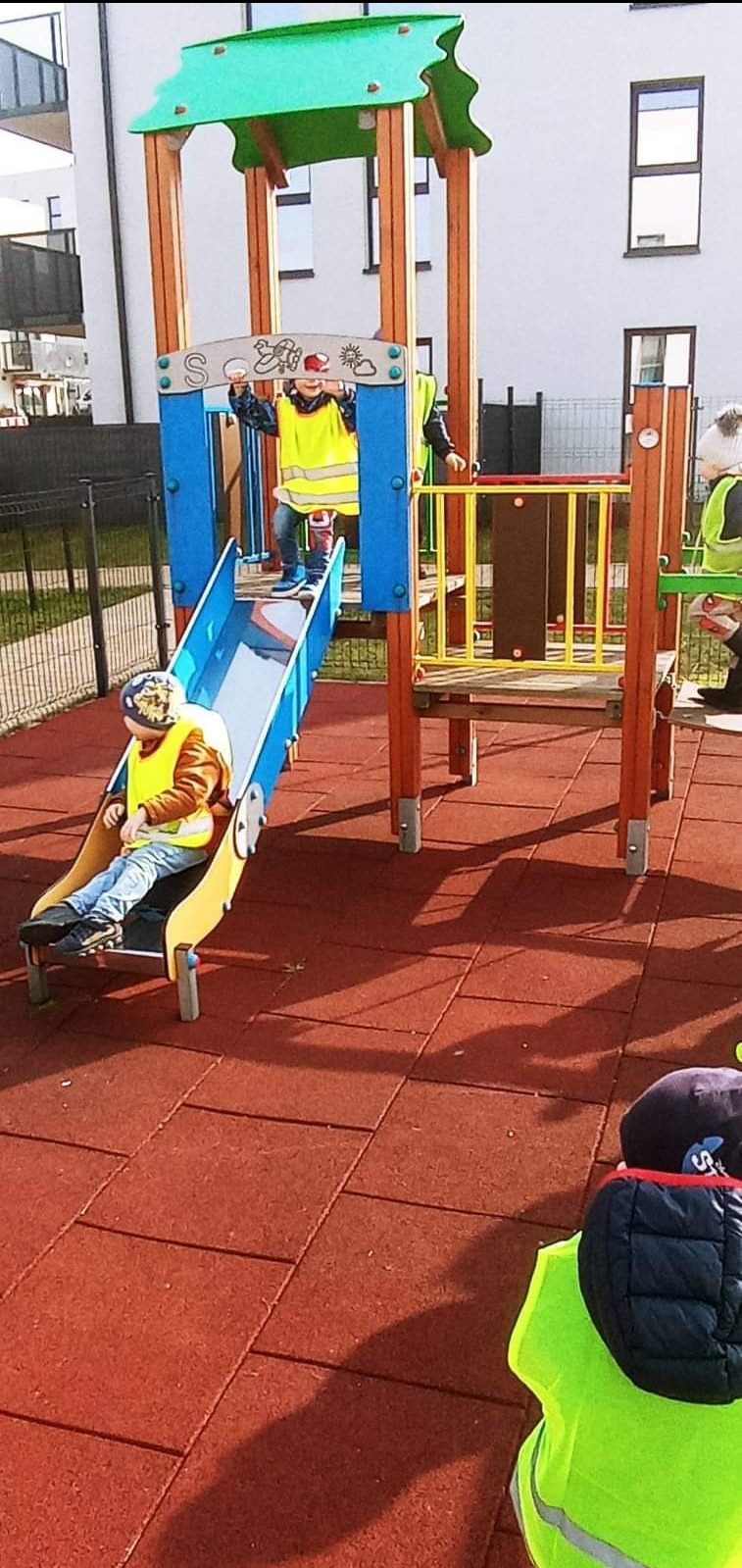Dzieci w jaskrawych kamizelkach bawią się na kolorowym placu zabaw z drewnianą konstrukcją, zjeżdżalnią i platformą, na tle bloku mieszkalnego.