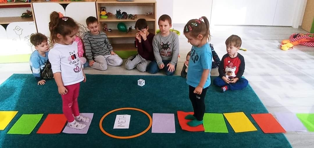 Dzieci bawią się w grę planszową z kolorowymi polami na dywanie, rzucają kostką i przechodzą przez obręcz z napisem META.