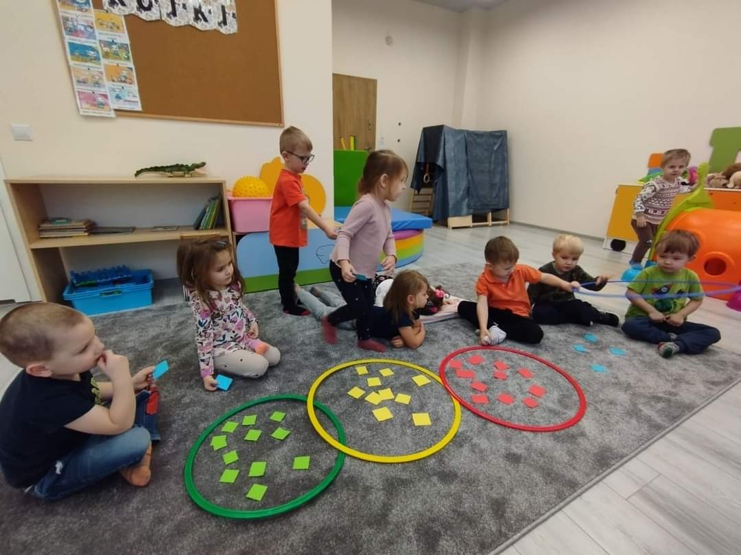 Grupa dzieci w sali bawi się kolorowymi obręczami i kwadratowymi kartami, siedząc na szarym dywanie; widoczny regał z zabawkami i dekoracje ścienne.