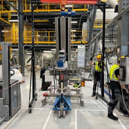 Industrial Service - Dwóch pracowników w żółtych kamizelkach i kaskach, montuje lub naprawia maszynę przemysłową za pomocą wózka widłowego i suwnicy w hali produkcyjnej z żółtymi elementami konstrukcyjnymi. Widoczne...