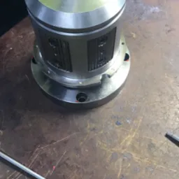 Metalowa obudowa cylindryczna z otworami, dokręcana kluczem imbusowym nasadowym na stole roboczym z widocznymi śladami użytkowania, w tle ręka w niebieskiej rękawicy roboczej i fragment odzieży...