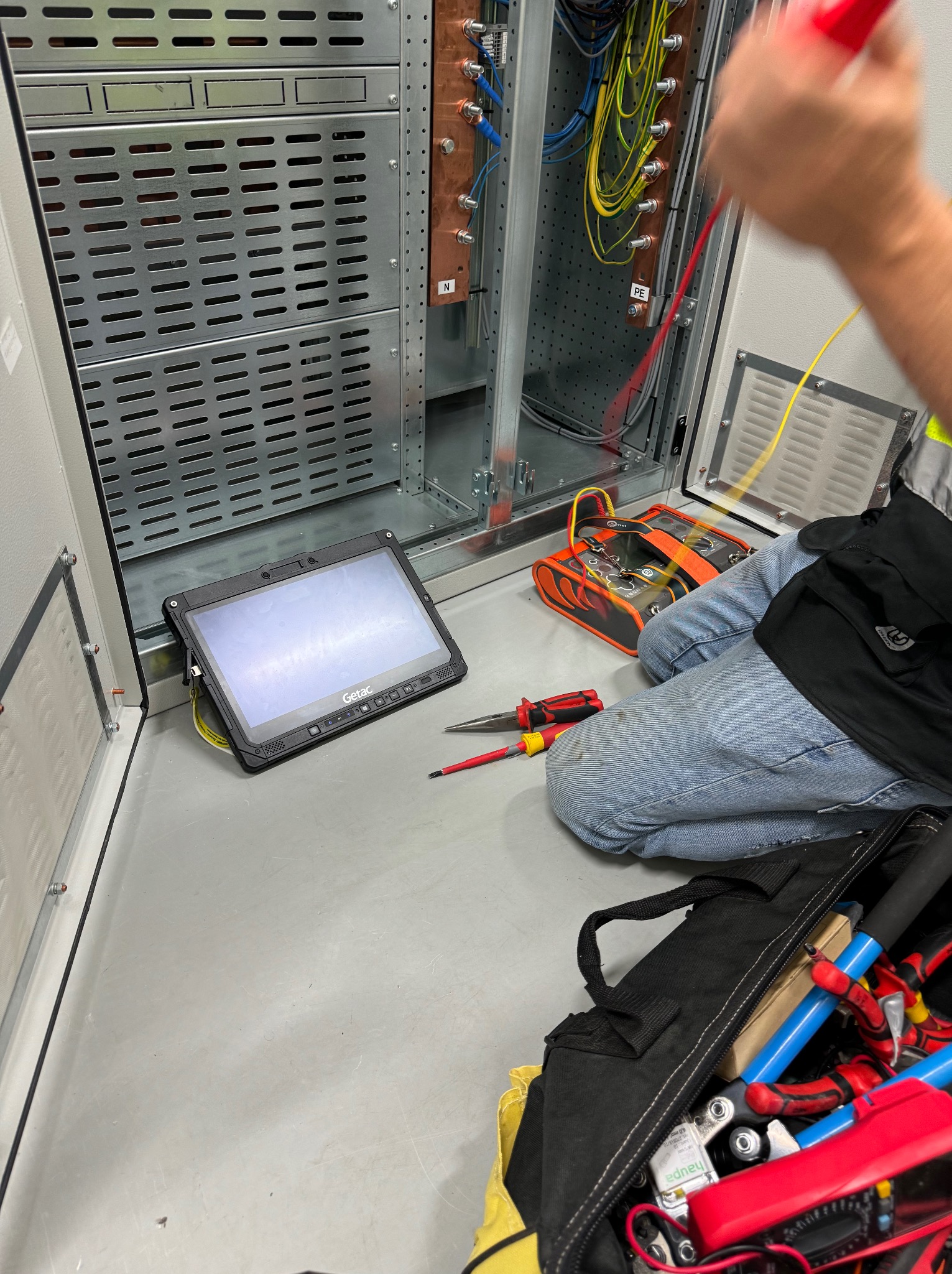 Elektryk w Legnicy wykonuje pomiary w rozdzielnicy elektrycznej, używając miernika i tabletu Getac. Widoczna skrzynka z narzędziami i przewody.