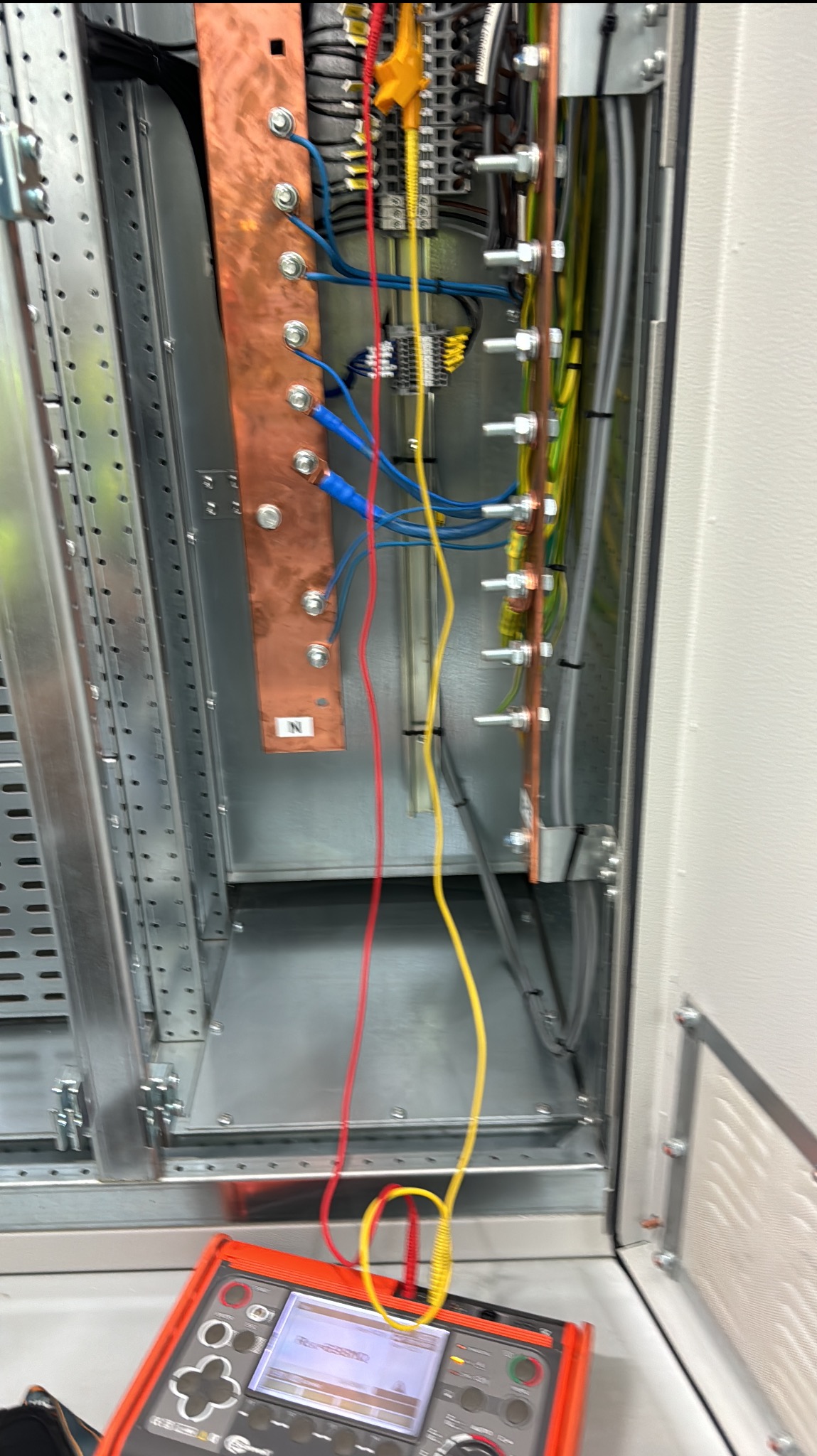Panel elektryczny w trakcie pomiarów, widoczne miedziane szyny, kable w różnych kolorach podłączone do listwy zaciskowej oraz miernik elektryczny z wyświetlaczem.