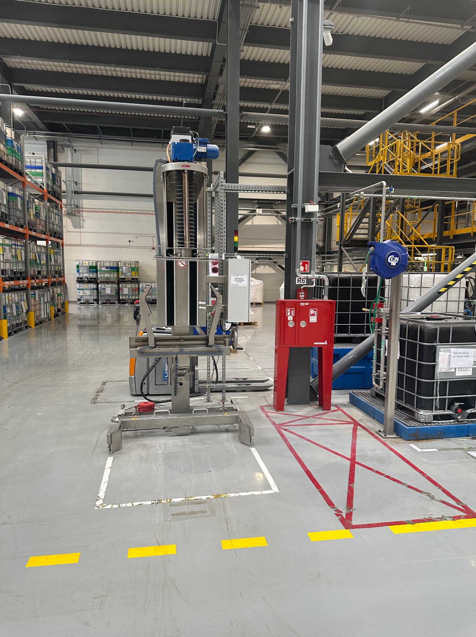 Wnętrze hali produkcyjnej z urządzeniem do podnoszenia i transportu pojemników, widoczne regały z pojemnikami w tle, oznakowanie podłogi w postaci żółtych i czerwonych linii.