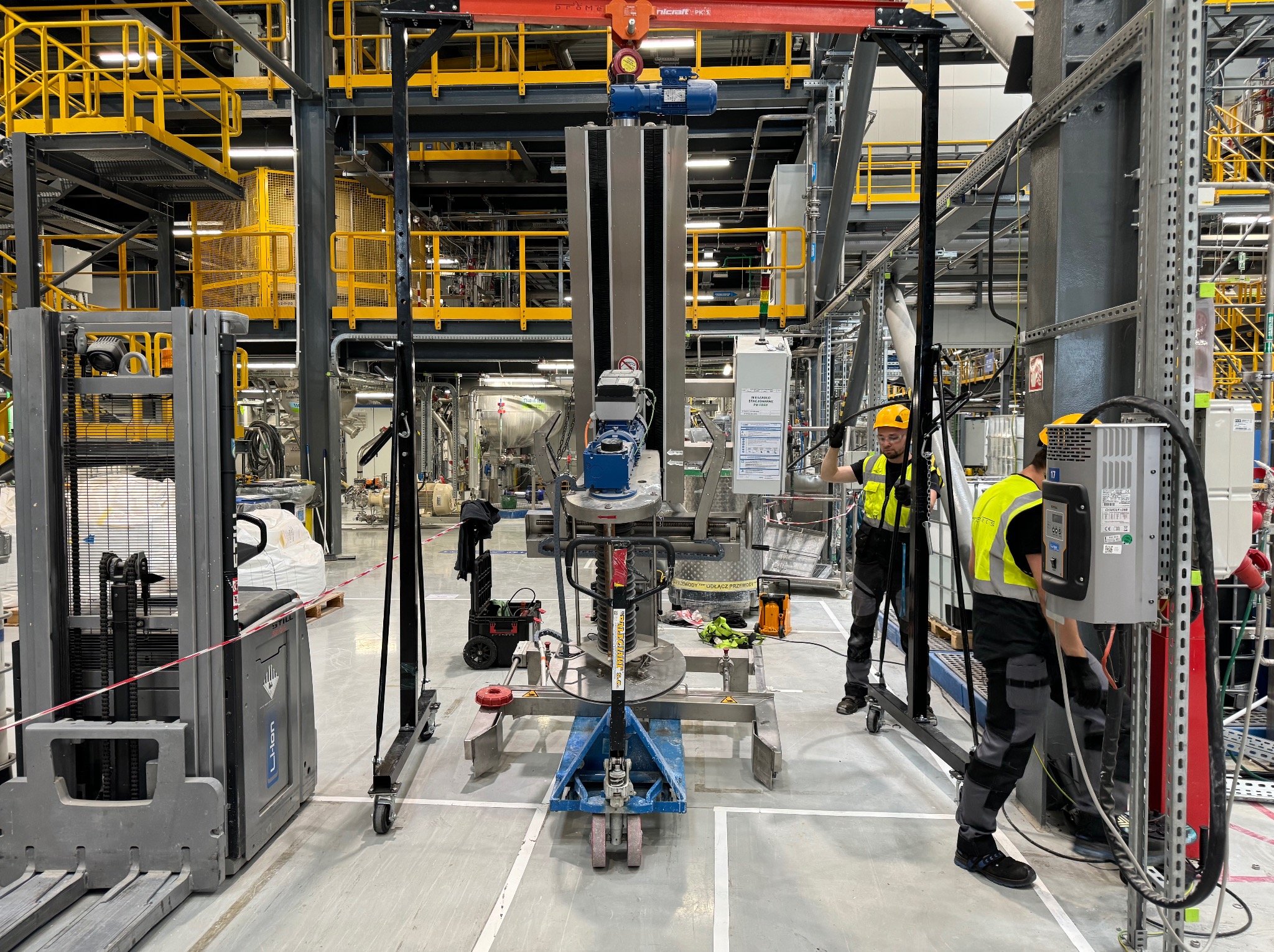 Dwóch pracowników w żółtych kamizelkach i kaskach, montuje lub naprawia maszynę przemysłową za pomocą wózka widłowego i suwnicy w hali produkcyjnej z żółtymi elementami konstrukcyjnymi. Widoczne...