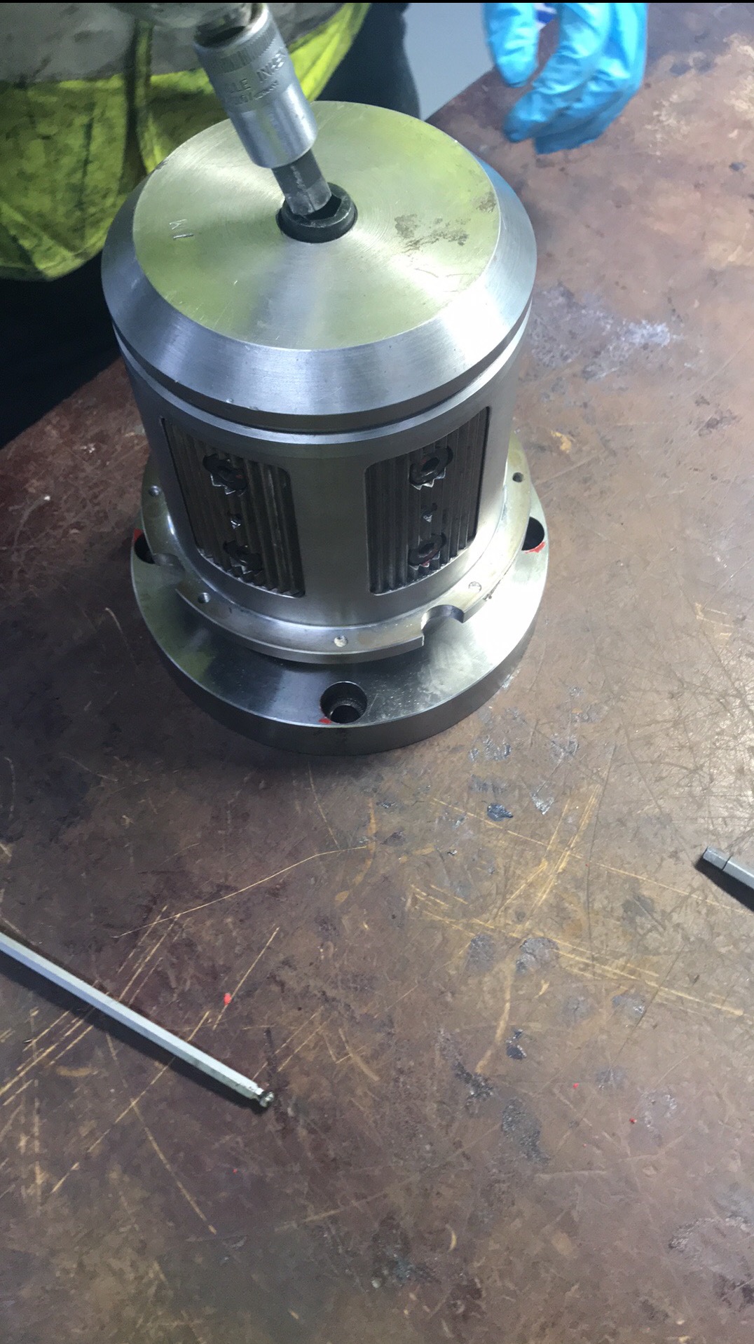 Metalowa obudowa cylindryczna z otworami, dokręcana kluczem imbusowym nasadowym na stole roboczym z widocznymi śladami użytkowania, w tle ręka w niebieskiej rękawicy roboczej i fragment odzieży...