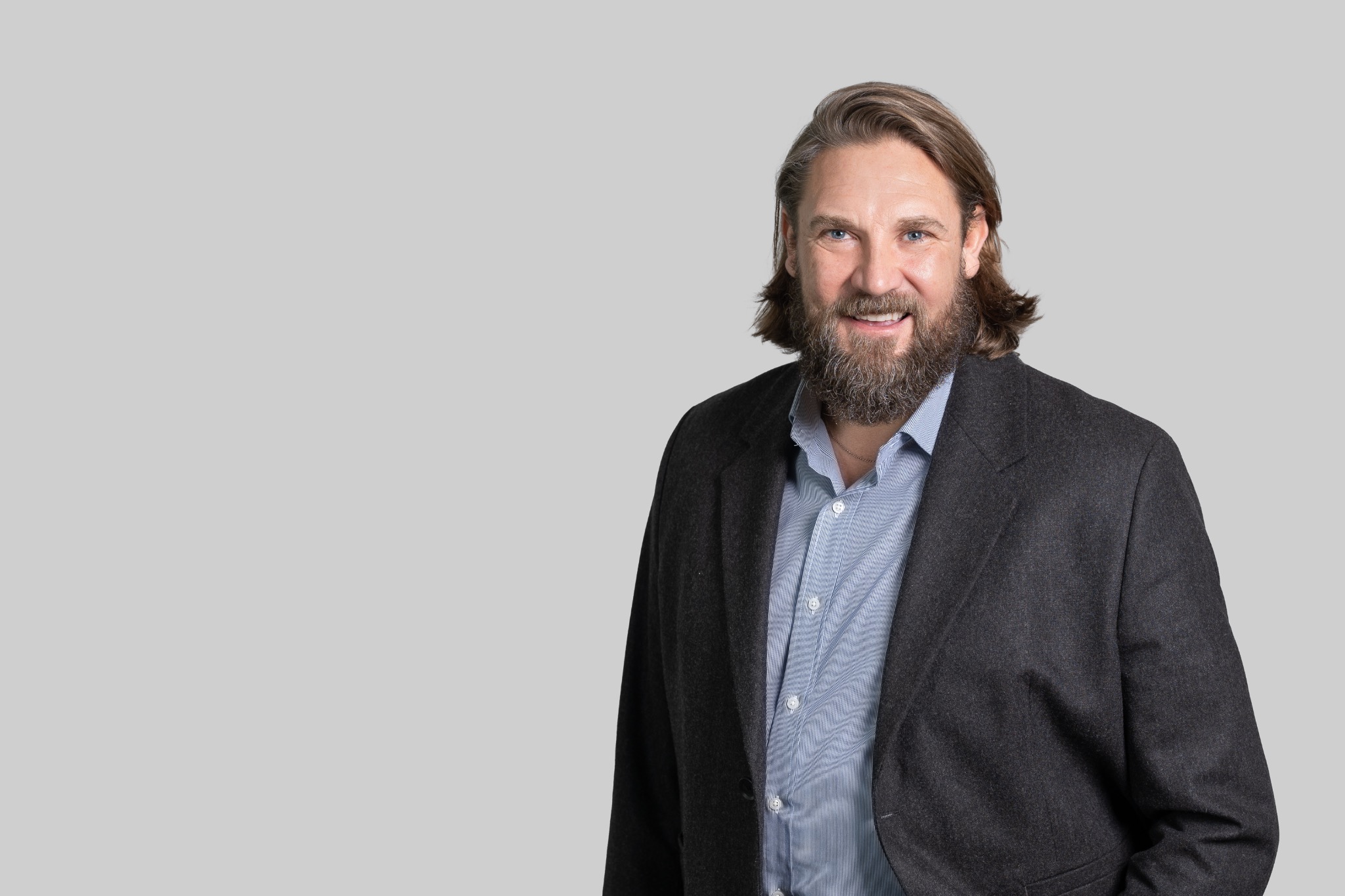 Portret uśmiechniętego mężczyzny z brodą i długimi włosami w szarej marynarce i niebieskiej koszuli na jasnoszarym tle.