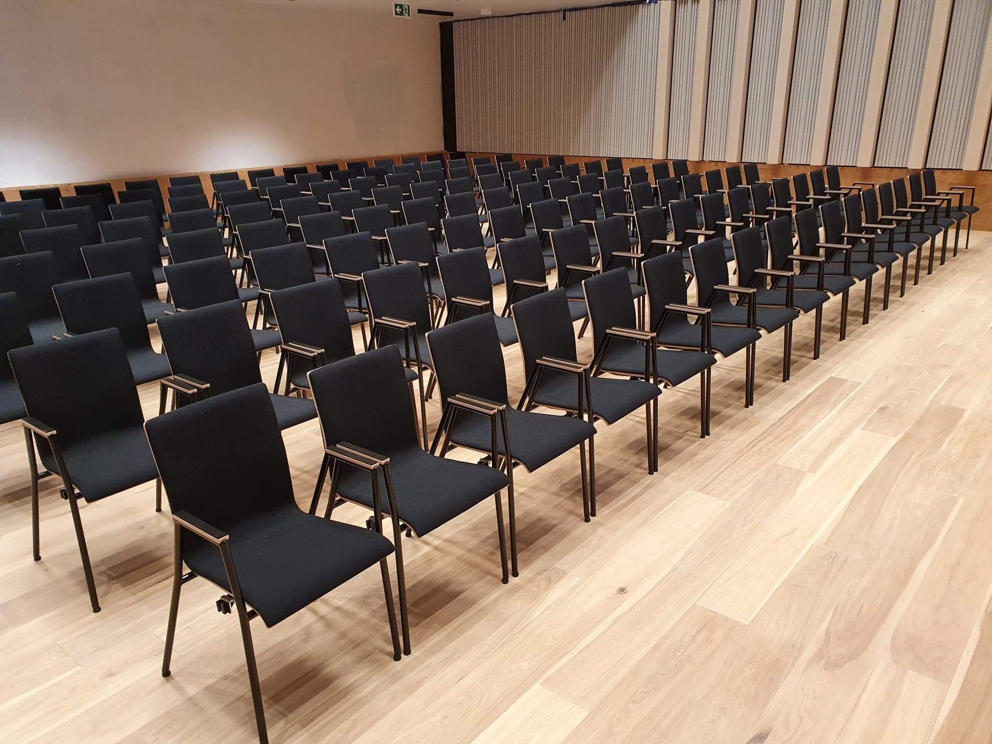 Rząd krzeseł konferencyjnych z czarnymi siedziskami i oparciami oraz metalowymi ramami, ustawionych na jasnej drewnianej podłodze w sali konferencyjnej z panelami ściennymi w tle.