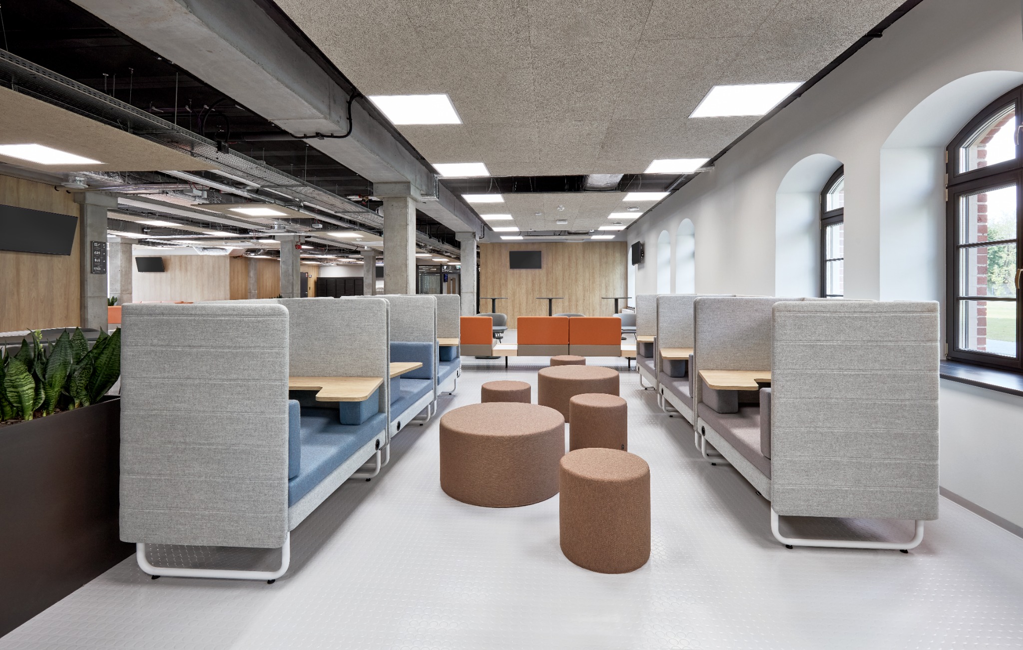 Nowoczesna przestrzeń biurowa z szarymi modułowymi sofami z drewnianymi stolikami, brązowymi pufami i jasnymi oknami łukowymi.