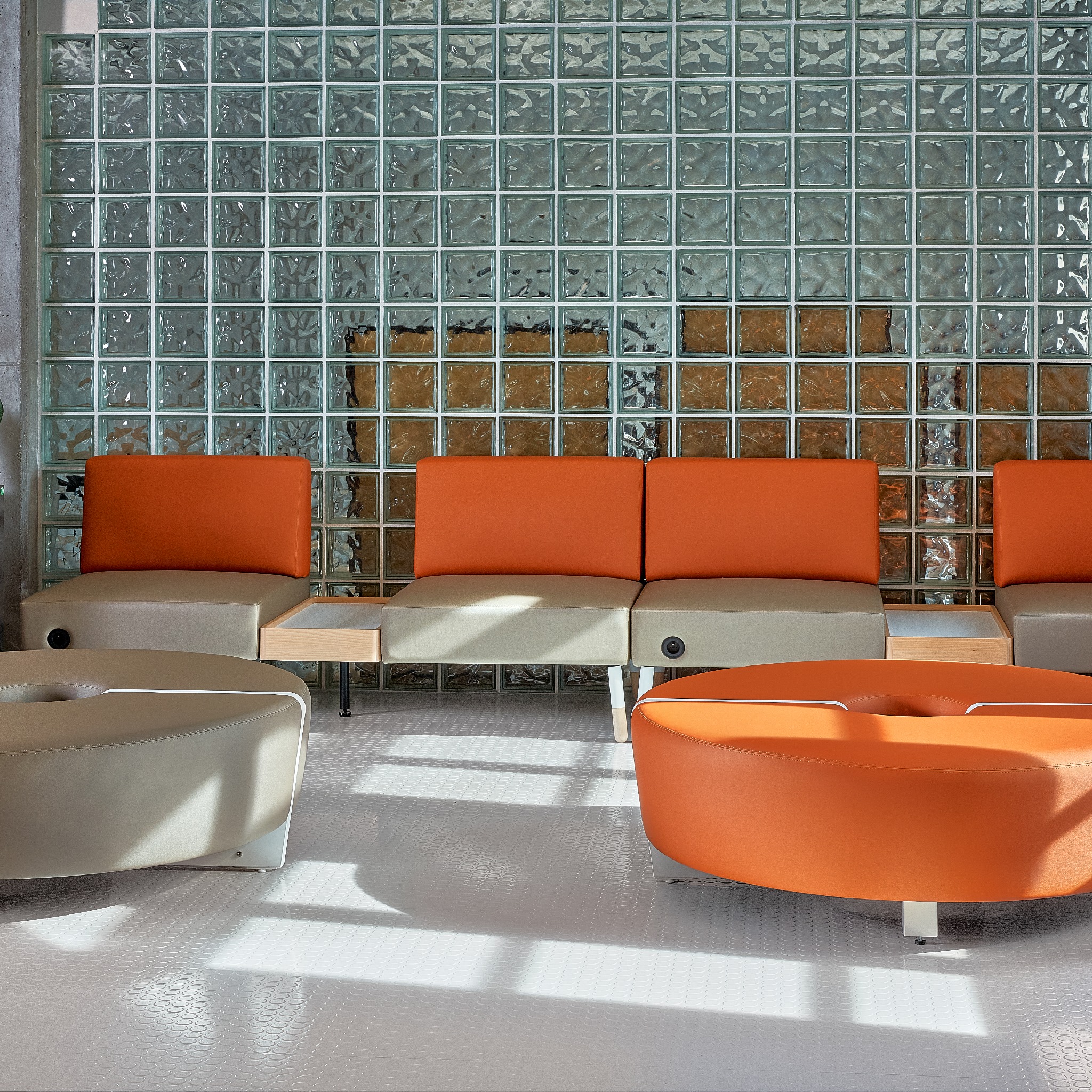 Nowoczesna poczekalnia z modułową sofą w odcieniach beżu i pomarańczy, stoliki boczne z jasnego drewna, ściana z luksferów, wzorzysta podłoga w jasnych barwach.