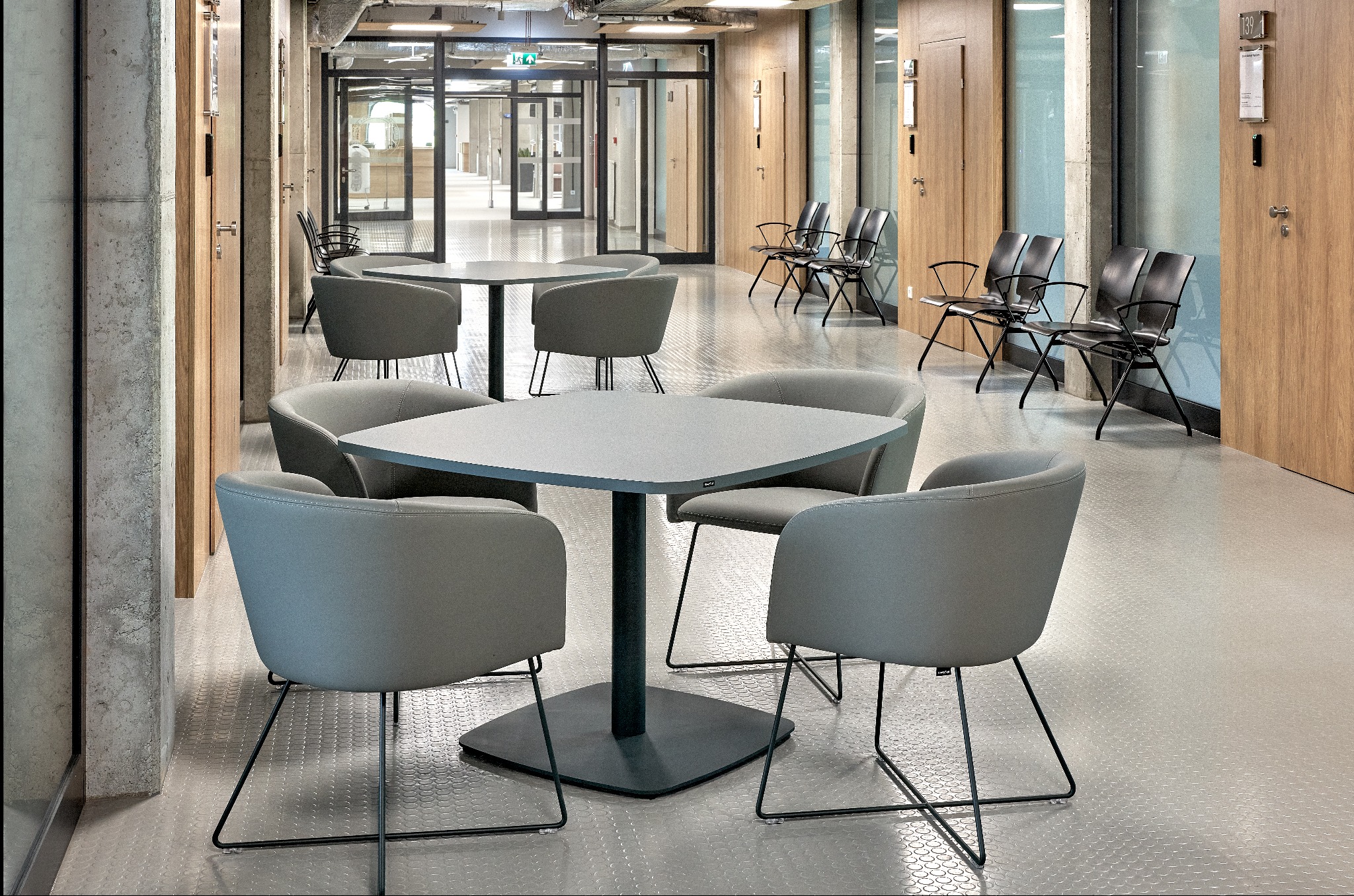 Nowoczesna przestrzeń biurowa z szarymi krzesłami i stolikami na metalowych nogach, betonowe kolumny i drewniane drzwi w tle, minimalistyczny design