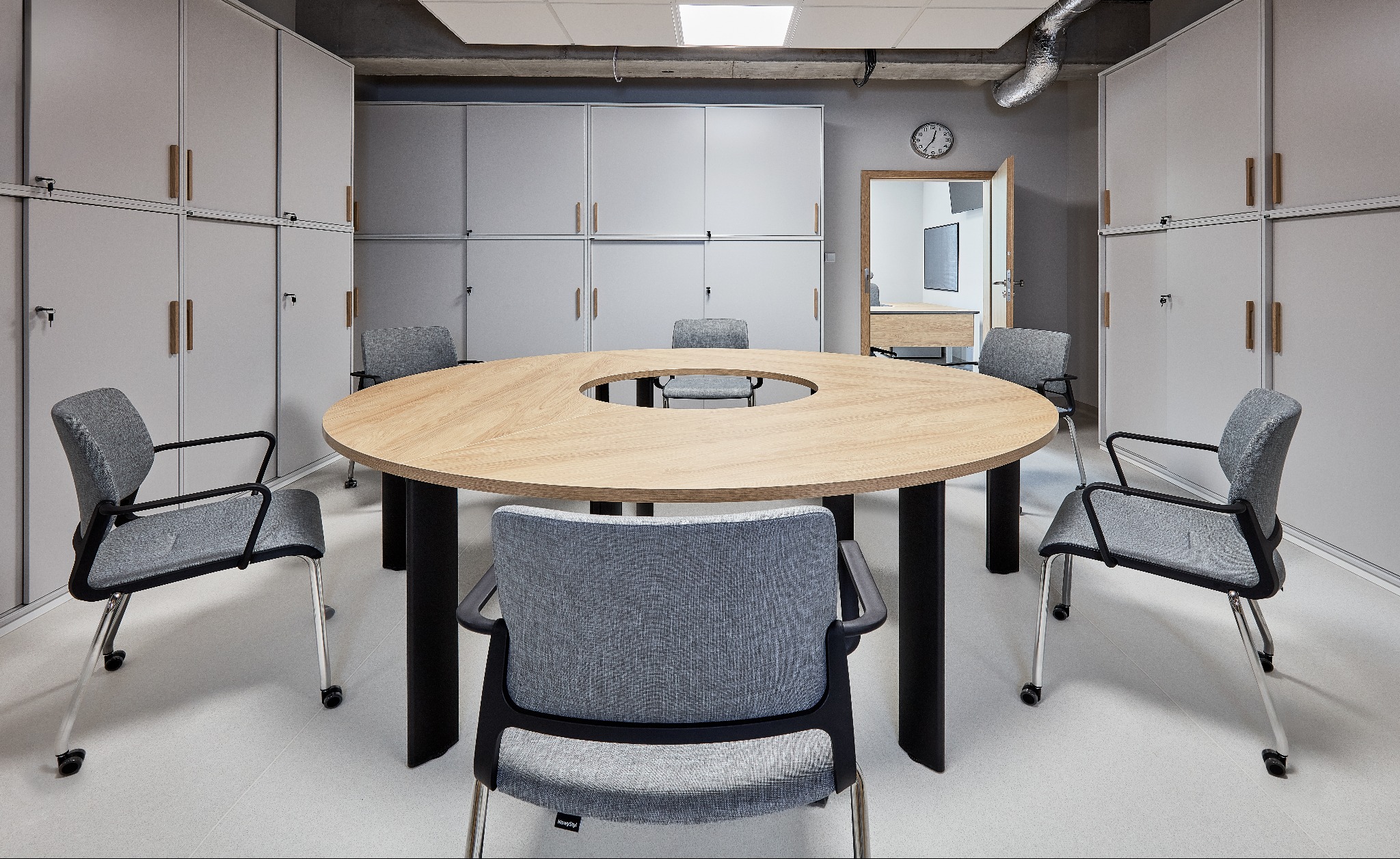 Jasne biuro z okrągłym stołem konferencyjnym z otworem w środku, otoczonym szarymi krzesłami na kółkach i szafami w tle.