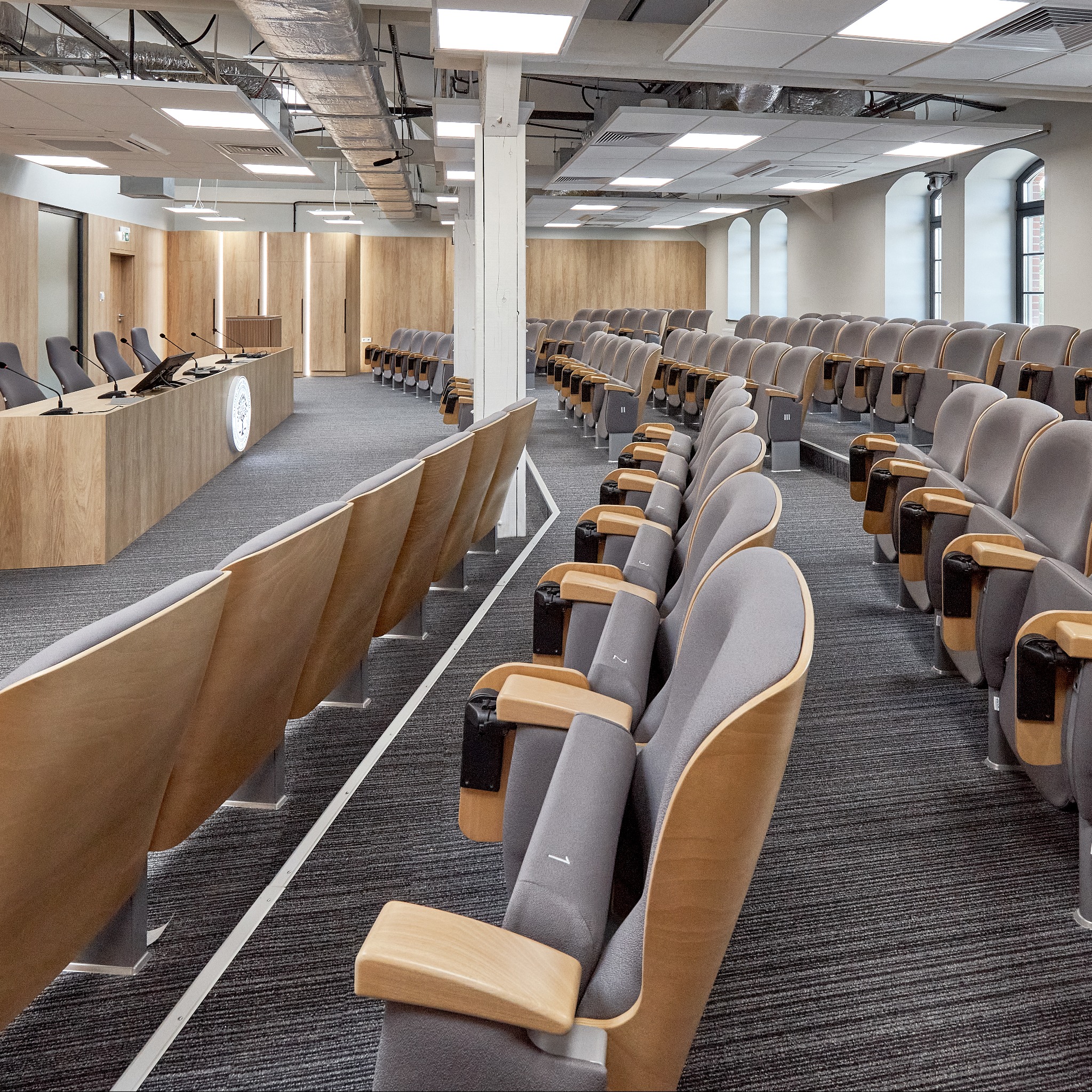 Sala konferencyjna z rzędami drewnianych krzeseł z szarymi siedziskami, mikrofonami na drewnianym podwyższeniu i oświetleniem sufitowym.