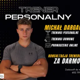 Młody mężczyzna, Michał Dargacz, z założonymi rękami na tle plakatu reklamującego treningi personalne, domowe i online, konsultacje trenerskie za darmo. Na nadgarstku widoczny smartwatch.